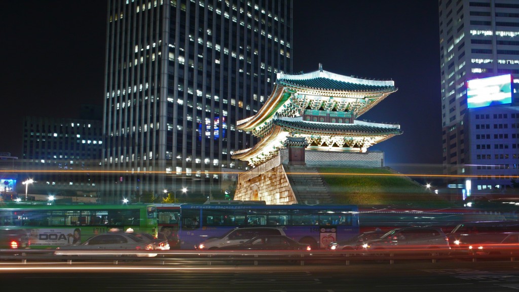 Long exposure photograph of cars passing in front of an historical building with a modern building in the background at night.